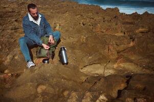 männlich Tourist mit Rucksack und Thermosflasche Flasche, Sitzung auf das Cliff durch Meer, genießen seine Kaffee brechen auf das Natur foto