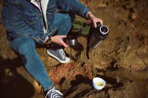 Nahansicht männlich Tourist Reisender Gießen Tee von ein touristisch Thermosflasche in ein Tasse, Sitzung auf das felsig Cliff im durch Ozean foto