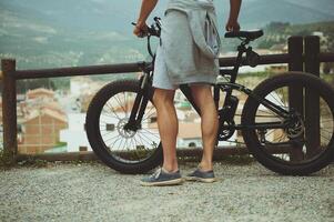 Nahansicht Aussicht von das zurück zu ein jung aktiv Mann Radfahrer Stehen in der Nähe von seine elektrisch Batterie angetrieben Fahrrad, bewundern schön Berge, genießen Reiten im das Natur. Personen. Öko Transport. Freiheit. foto