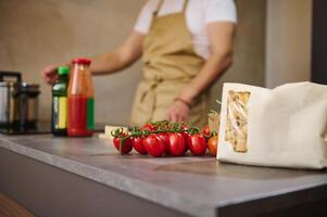 selektiv Fokus auf ein Bündel von rot reif organisch Tomate Kirsche und ein Papier Paket mit Pasta, gegen männlich Koch Stehen beim Induktion Herd, einstellen Programm, Kochen Abendessen im modern Küche Innere foto