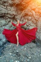 rot Kleid Felsen Frau. ein blond mit fließend Haar im ein lange fließend rot Kleid steht in der Nähe von ein Felsen von vulkanisch Herkunft. Reise Konzept, Foto Session beim Meer