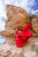 ein Frau im ein rot Seide Kleid steht durch das Ozean, mit Berge im das Hintergrund, wie ihr Kleid schwankt im das Brise. foto