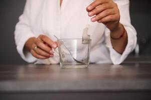 Nahansicht Frau Hände halten ein Tee Tasche Über ein leeren Glas Tasse im das Zuhause Küche, vorbereiten heiß trinken zum Frühstück im das Morgen foto