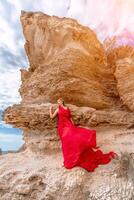 Frau rot Seide Kleid steht durch das Ozean, mit Berge im das Hintergrund, wie ihr Kleid schwankt im das Brise. foto