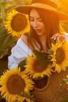 ein Mädchen im ein Hut auf ein schön Feld von Sonnenblumen gegen das Himmel im das Abend Licht von ein Sommer- Sonnenuntergang. Sonnenstrahlen durch das Blume Feld. natürlich Hintergrund. foto