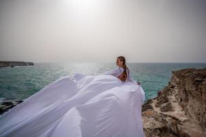 glücklich Freiheit Frau auf das Strand genießen und posieren im Weiß Kleid. Rückseite Aussicht von ein Mädchen im ein flattern Weiß Kleid im das Wind. Feiertage, Ferien beim Meer. foto