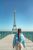 groß Modell- von das Eiffel Turm auf das Strand. ein Frau Spaziergänge entlang das Seebrücke gegenüber das Turm, tragen ein Blau Jacke und Weiß Jeans. foto