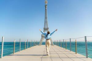 groß Modell- von das Eiffel Turm auf das Strand. ein Frau Spaziergänge entlang das Seebrücke gegenüber das Turm, tragen ein Blau Jacke und Weiß Jeans. foto