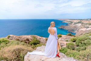 blond mit lange Haar auf ein sonnig Strand im ein Weiß fließend Kleid, Rückseite Sicht, Seide Stoff winken im das Wind. gegen das Hintergrund von das Blau Himmel und Berge auf das Strand. foto