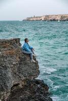 ein Frau im ein Blau Jacke sitzt auf ein Felsen über ein Cliff über das Meer, suchen beim das stürmisch Ozean. Mädchen Reisender ruht, denkt, Träume, genießt Natur. Frieden und Ruhe Landschaft, windig Wetter. foto