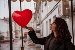 ein Frau halten ein rot Herz Ballon. das Frau ist tragen ein schwarz Mantel und hat rot Haar. das Szene ist einstellen im ein Stadt Straße mit Autos und ein Gebäude im das Hintergrund. foto