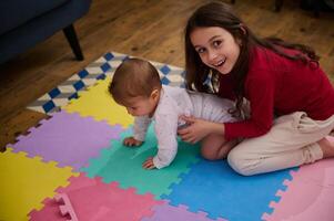 bezaubernd wenig Mädchen lächelnd suchen beim Kamera, ausdrücken positiv Emotionen und Glück Portion ihr Bruder, ein süß Baby Junge auf kriechen auf bunt Puzzle Teppich beim heim. Kinder. Wachstum. Familie foto