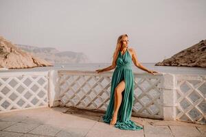 Frau Meer reise Grün Kleid. Seite Aussicht ein glücklich Frau mit lange Haar im ein lange Minze Kleid posieren auf ein Strand mit Ruhe Meer Bokeh Beleuchtung auf sonnig Tag. Mädchen auf das Natur auf Blau Himmel Hintergrund. foto