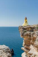 Frau im ein Gelb Kleid auf das Meer. Seite Aussicht jung schön sinnlich Frau im Gelb lange Kleid posieren auf ein Felsen hoch über das Meer beim Sonnenuntergang. Mädchen im Natur gegen das Blau Himmel foto