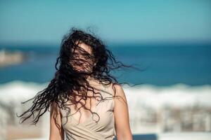 Meer Frau ausruhen. Porträt von ein Frau mit lange lockig schwarz Haar im ein Beige Kleid steht auf ein Balkon gegen das Hintergrund von das Meer. Tourist Ausflug zu das Meer. foto