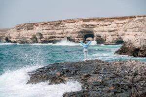 ein Frau im ein Blau Jacke steht auf ein Felsen über ein Cliff über das Meer und sieht aus beim das tobt Ozean. Mädchen Reisender ruht, denkt, Träume, genießt Natur. Frieden und Ruhe Landschaft, windig Wetter. foto