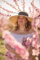 Frau Blühen Pfirsich Obstgarten. gegen das Hintergrund von ein malerisch Pfirsich Obstgarten, ein Frau im ein lange Weiß Kleid und Hut genießt ein friedlich gehen im das Park, umgeben durch das Schönheit von Natur. foto