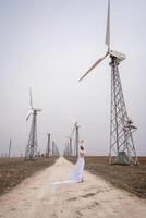 ein Frau im ein Weiß Kleid ist Gehen Nieder ein Schmutz Straße im Vorderseite von ein Reihe von Wind Turbinen. foto