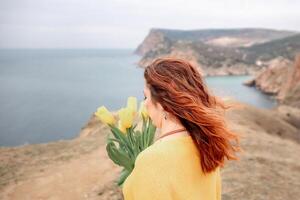 Rückseite Aussicht von ein Frau mit lange Haar gegen ein Hintergrund von Berge und Meer. halten ein Strauß von Gelb Tulpen im ihr Hände, tragen ein Gelb Sweatshirt foto
