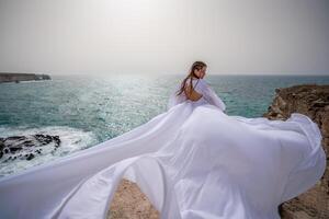 glücklich Freiheit Frau auf das Strand genießen und posieren im Weiß Kleid. Rückseite Aussicht von ein Mädchen im ein flattern Weiß Kleid im das Wind. Feiertage, Ferien beim Meer. foto