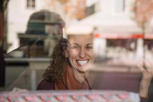 strahlend jung Frau lächelnd hinter ein reflektierend Fenster im ein städtisch Cafe Rahmen foto