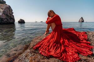 schön sinnlich Frau im ein fliegend rot Kleid und lange Haar, Sitzung auf ein Felsen über das schön Meer im ein groß Bucht. foto