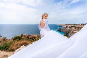 blond mit lange Haar auf ein sonnig Strand im ein Weiß fließend Kleid, Rückseite Sicht, Seide Stoff winken im das Wind. gegen das Hintergrund von das Blau Himmel und Berge auf das Strand. foto