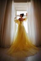 ein Frau Silhouette im ein golden luxuriös Kleid gegen das Hintergrund von ein Fenster hält ein Vorhang mit ihr Hände. elegant Dame im ein Gelb lange Seide Kleid mit nackt zurück, zurück Sicht. foto