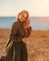 Porträt blond Meer Kap. ein Ruhe jung blond im ein aufgeknöpft khaki Regenjacke steht auf das Strand, unter das Regenjacke Dort ist ein schwarz Rock und oben foto