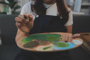 abgeschnitten Bild von weiblich Künstler Stehen im Vorderseite von ein Staffelei und Eintauchen Bürste in Farbe Palette foto