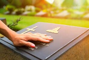 Frau Hand berühren das schwarz Stein Grab beim Friedhof mit Orange Sonnenlicht aufflackern. in Erinnerungen schwelgen, vermissen, traurig und verlieren Person im Familie oder wichtig Menschen foto