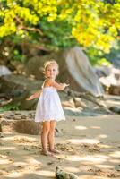 wenig Mädchen im Weiß Kleid haben Spaß auf Strand im das Schatten von Bäume und Palmen. foto