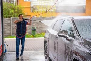 jung Mann wäscht seine Auto beim ein Selbstbedienung Auto waschen mit ein Schlauch mit Druck- Wasser und Schaum. hoch Qualität Foto