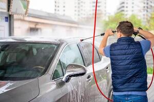 jung Mann wäscht seine Auto beim ein Selbstbedienung Auto waschen mit ein Schlauch mit Druck- Wasser und Schaum. hoch Qualität Foto