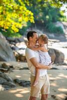 wenig Mädchen auf Vaters Hände Lächeln und jubeln auf Strand im das Schatten von Bäume und Palmen. glücklich Familie Konzept foto