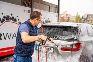 Prag, Tschechisch Republik - - April 5, 2024. jung Mann wäscht seine Auto beim ein Selbstbedienung Auto waschen mit ein Schlauch mit Druck- Wasser und Schaum. hoch Qualität Foto