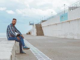 Bärtiger Hipster-Mann mit Mohawk sitzt auf einer Betonbank am Meer am Wasser. foto