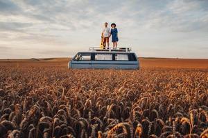 Paar Mann mit Gitarre und Frau mit Hut stehen auf dem Dach eines Autos in einem Weizenfeld foto