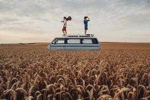 Paar Mann mit Gitarre und Frau mit Hut stehen auf dem Dach eines Autos in einem Weizenfeld foto