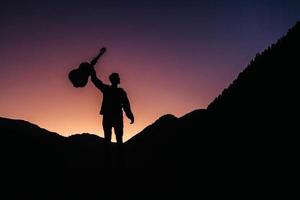 Silhouette eines Mannes, der eine Gitarre vor dem Hintergrund von Bergen und Sonnenuntergang hält foto