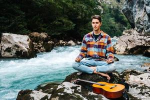 Mann in meditativer Position mit Gitarre sitzt am Ufer eines Bergflusses auf einem Hintergrund von Felsen und Wald foto