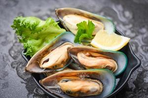 frische Muscheln auf schwarzer Schüssel und Eis mit Zitrone und Salat für gekochtes Essen - Meeresfrüchte, Schalentiere, gedämpfte Muscheln
