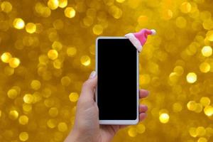 Mock-up-Telefon mit Weihnachtsmütze in der Hand der Frau auf goldenem Hintergrund mit Boke. Weihnachts-Online-Verkaufskonzept foto