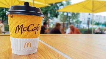 Mccafe-Menü im Mcdonalds Restaurant. Gelbe Tasse Kaffee auf einem Tisch im Freien mit gelben Regenschirmen und Besuchern im Hintergrund. Frühstück, Kaffeezeit. Ukraine, Kiew - 6. September 2019. foto