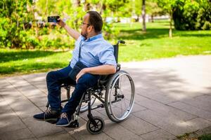 Porträt von glücklich Mann im Rollstuhl. er ist genießen sonnig Tag im Stadt Park und nehmen Selfie. foto