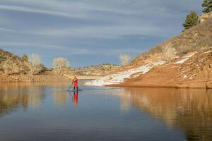 männlich Paddler im rot Trockenanzug ist Paddeln ein Stand oben Paddel Boot auf Berg See im Colorado, Winter Landschaft foto