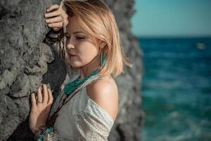 Mitte alt Frau sieht aus gut mit blond Haar, Boho Stil im Weiß lange Kleid auf das Strand Dekorationen auf ihr Hals und Waffen. foto