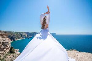 blond mit lange Haar auf ein sonnig Strand im ein Weiß fließend Kleid, Rückseite Sicht, Seide Stoff winken im das Wind. gegen das Hintergrund von das Blau Himmel und Berge auf das Strand. foto