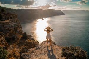 ein Frau steht auf ein Cliff mit Blick auf das Ozean. sie ist tragen ein Weiß Kleid und ein Stroh Hut. das Himmel ist wolkig und das Sonne ist Einstellung. foto