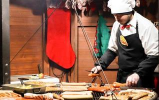 Ukraine, Kiew - 3. Januar 2019. Ein Koch kocht ein Barbecue auf dem Grill, Street Food, Food Festival. frische Wurst und gegrillte Hotdogs im Freien. ein Mann bereitet Streetfood der deutschen Küche zu foto